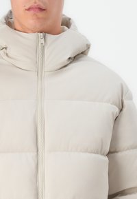 Beige színű pufi dzseki kapucnival, elöl cipzáros, steppelt mintázattal. Az anyag lágy és könnyűnek tűnik.