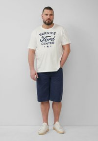 Witte katoenen T-shirt met marineblauwe bedrukte tekst "SERVICE FORD CENTER", gecombineerd met marineblauwe shorts en witte sneakers met lichtbruine zolen.