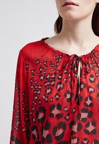 Blusa rossa trasparente con scollo arricciato e macchie leopardate nere. Dettaglio con cordoncino al collo, maniche fluide e texture leggera.