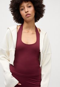 Top halter em bordeaux com decote redondo e hoodie cropped branco com fecho. Textura suave e ajuste descontraído, destacando a pele por baixo.