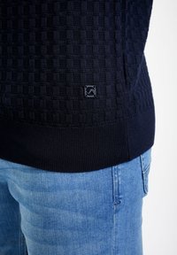 Gros plan d'une personne portant un pull texturé bleu marine foncé avec un petit patch logo au-dessus de l'ourlet côtelé et un jean en denim bleu clair.