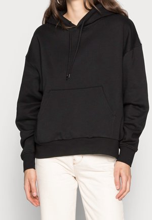 Femme portant un sweat à capuche noir avec poche avant et cordons, associée à un pantalon blanc cassé, debout devant un fond uni.