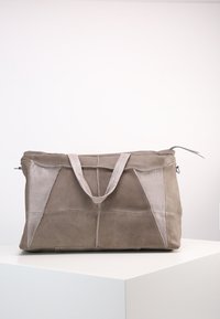 Sac de week-end en daim et cuir marron doux, avec des poignées doubles, reposant sur un piédestal blanc contre un fond uni.