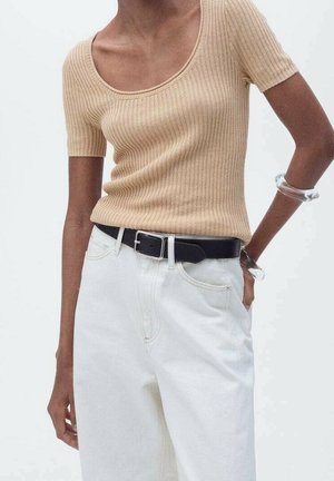 Femme portant un haut beige côtelé à manches courtes rentré dans un pantalon blanc taille haute avec une ceinture noire et des bracelets transparents au poignet.