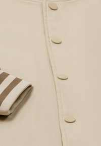 Top din țesătură bej cu cinci butoane mari bej; manșete tricotate în dungi de crem și brun, evidențiind textura netedă și designul elegant.