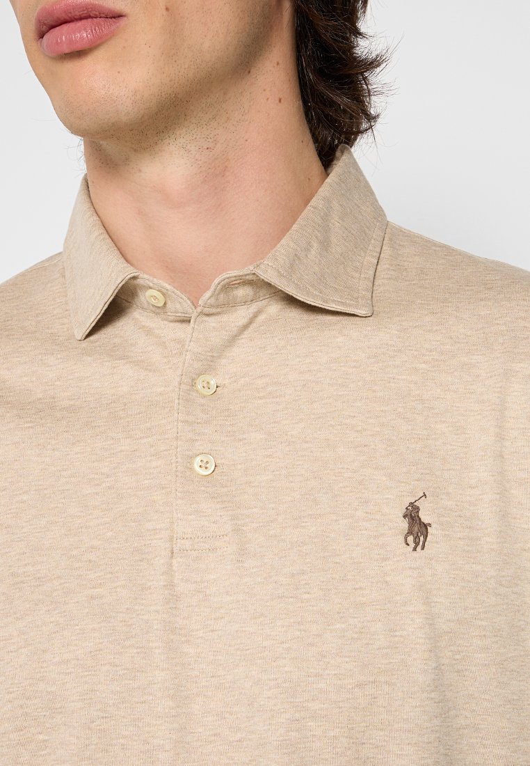 Polo beige en tissu doux, avec un col classique, trois boutons et un logo brodé marron sur la poitrine gauche.