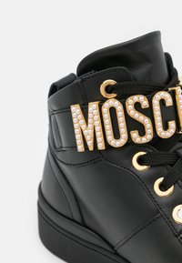 Svart högsneaker med gyllene öglor och dekorativa "MOSCH"-bokstäver prydda med små vita pärlor på svarta snören.