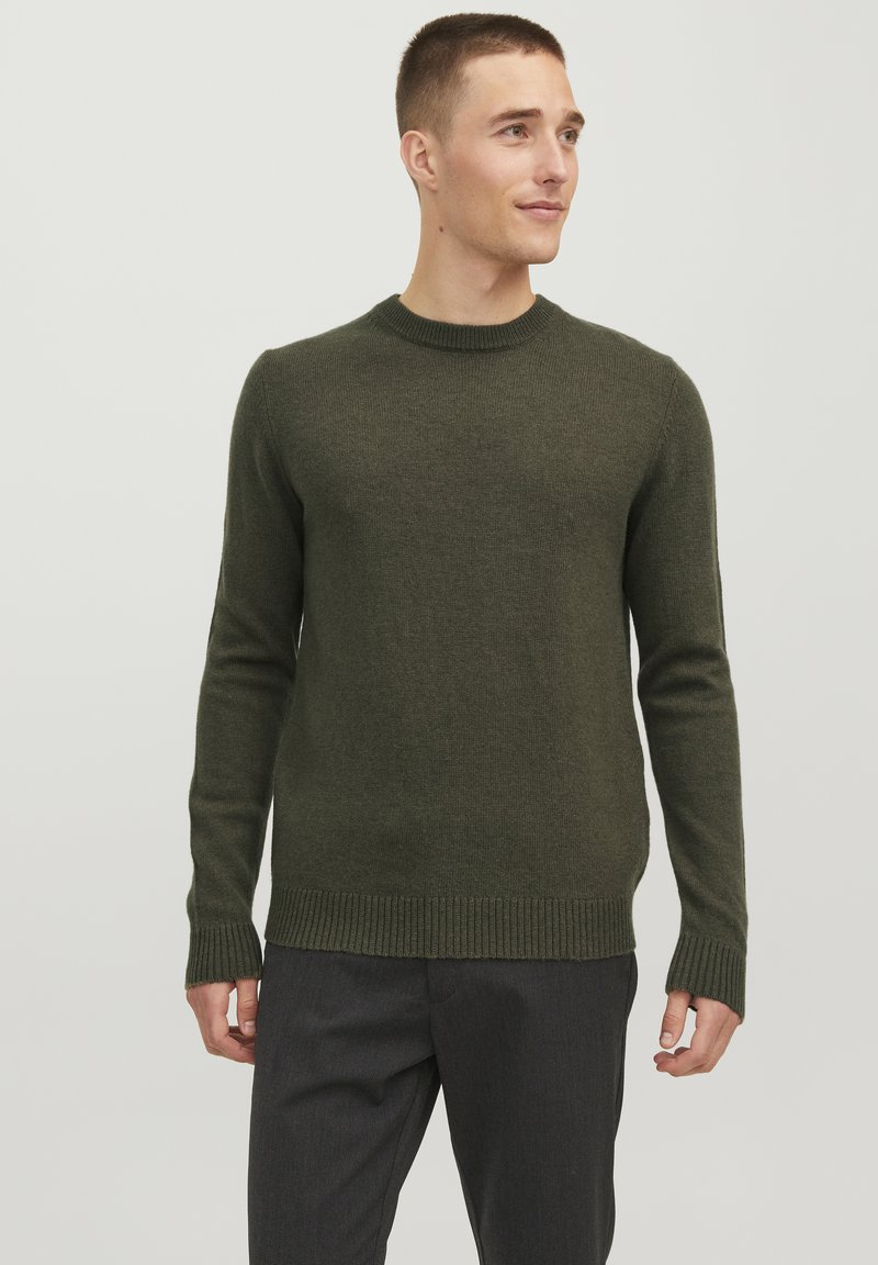 Pull à col rond en tricot vert olive, avec des poignets et un ourlet côtelés. La texture du tissu est douce et légèrement ajustée. Associé à un pantalon sombre à motifs.