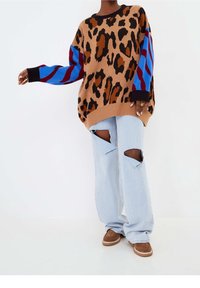 Pull surdimensionné imprimé léopard marron avec manches rayées rouges et bleues, associé à un jean bleu clair déchiré et des chaussures marron.
