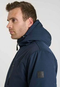 Giacca imbottita blu navy con cappuccio, dotata di tasca con zip e tessuto testurizzato. Include una toppa con logo sul maniche sinistro.