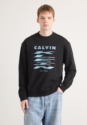 Fekete pulóver világoskék grafikával, amelyen a "CALVIN" felirat található, bordázott kerek nyakkal és hosszú ujjakkal. Világos farmernadrággal párosítva.