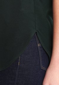 Tmavě zelená košile s bočním rozparkem, nošená přes tmavě modré džíny s viditelným žlutým prošíváním a částí nositelovy ruky.