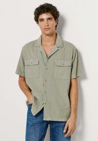 Młody mężczyzna o kręconych włosach, ubrany w jasnozieloną, krótkorękawową koszulę zapinaną na guziki z dwoma kieszeniami na piersi oraz niebieskie dżinsy, stoi na tle jasnej ściany.