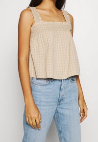 Top corto beige arricciato con spalline arricciate, caratterizzato da un motivo a scacchi. Abbinato a jeans azzurri chiari. Il tessuto è leggero e testurizzato.