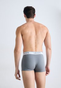 Boxer gris avec une taille blanche ornée du logo Lacoste. Le tissu semble lisse, épousant étroitement le corps.