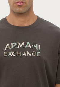 T-shirt in cotone grigio scuro con la scritta "ARMANI EXCHANGE" ricamata in lettere multicolori e texture, che mostra variazioni tonali e dimensione.