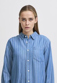 Camicia di jeans con bottoni caratterizzata da righe verticali bianche, colletto a punta, maniche lunghe e taschino sul petto. Il colore è azzurro chiaro.