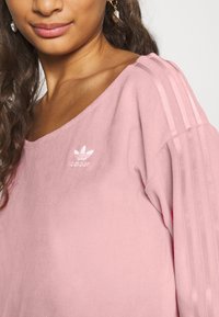 Chemise surdimensionnée rose en tissu doux. Présente un large décolleté et trois bandes blanches sur chaque épaule, avec le logo Adidas sur la poitrine.