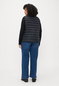 Juodas pūkuotas liemėnis su horizontaliu siuvinėjimu, dėvimas ant ilgomis rankovėmis marškinėlių, suderintas su plačiomis mėlynomis džinsinėmis kelnėmis ir masyviomis juodomis avalynėmis.