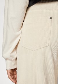 Beige corduroy broek met brede ribbels en een achterzak met een driehoekige flap. Zachte textuur met metalen knopenaccenten.