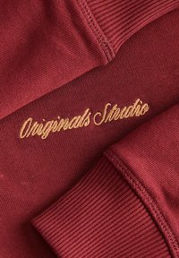 Maroon kangas muodokkaalla tekstuurilla, jossa on kultaiset brodeeraukset, joissa lukee "Originals Studio" ja ribattuja hihansuita kontrastina.