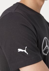 Czarna bawełniana koszulka z białym logo Pumy na lewym rękawie oraz grafiką na klatce piersiowej przypominającą emblemat samochodu.