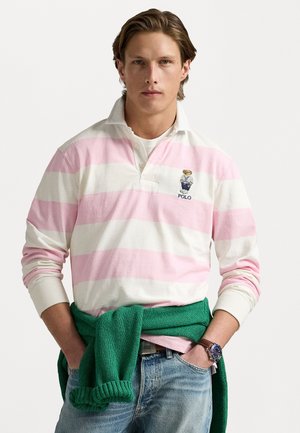 Langærmet polo-shirt i hvid med pink vandrette striber. Har en krave, tre-knaps placket og broderet logo. Bæres med en grøn sweater.