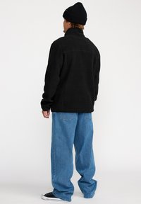 Svart fleecejacka med hög krage, kombinerad med ljusblå löst sittande jeans och svarta sneakers. En mössa fulländar outfiten.