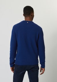 Gebreide blauwe trui met een gestructureerd patroon, ronde halslijn en ribgebreide manchetten en onderkant. Heeft een klein bruin logo op de achterkant.