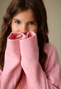 Maglione rosa lavorato a maglia con polsini a coste spessi e una finitura morbida e strutturata, che copre parzialmente mani e viso, con chiusura a bottoni.