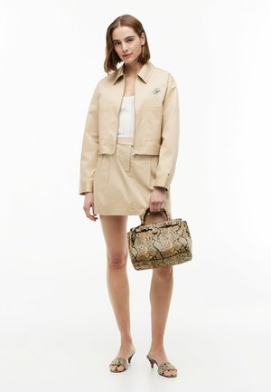 Kvinde iført beige jakke og nederdel, hvid top, der holder en håndtaske med slangeskinds mønster og iført matchende slangeskinds hæle.