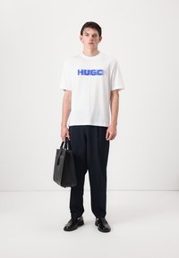 Hvid T-shirt med korte ærmer med blå "HUGO" tekst, sorte bukser, sort taske og skinnende sorte sko mod en ensfarvet baggrund.