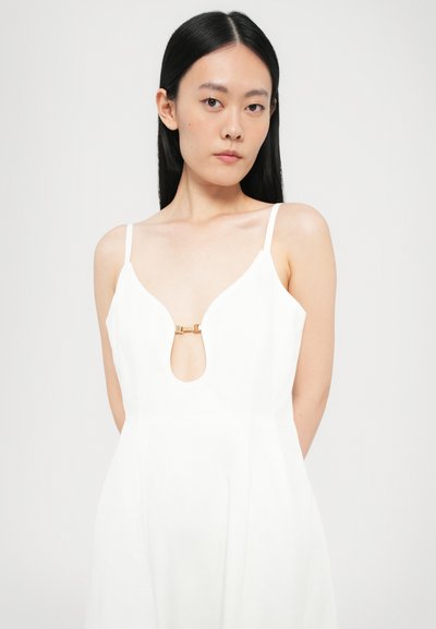 Vestido blanco con tirantes finos ajustables, que presenta un escote en V profundo y un detalle de ojal en la parte delantera, acentuado por un pequeño broche dorado.