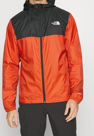 Mężczyzna noszący czerwoną i czarną kurtkę przeciwwiatrową z kapturem, z zamkiem z przodu i logo The North Face na piersi.
