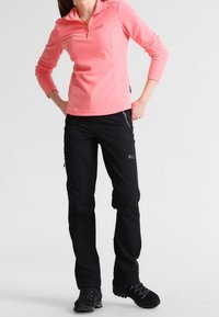 Jasnoróżowy pulower z ćwiartkowym zamkiem błyskawicznym i czarnymi spodniami outdoorowymi z bocznymi kieszeniami. Widoczne fakturowana tkanina i solidne rzemiosło.