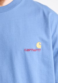 Lichtblauw katoenen t-shirt met een rood "carhartt" logo en een geel embleemontwerp op de linkerborst. Eenvoudige ronde hals.