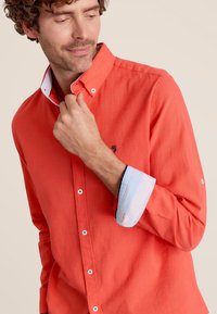 Chemise boutonnée corail avec un col et des poignets rayés bleu et blanc en contraste. Tissu léger avec une texture visible et un léger éclat.