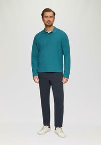 Türkises Langarm-Polohemd mit Kragen, kombiniert mit dunkelblauen Hosen und weißen Sneakers, mit dezentem Branding auf der Brust.