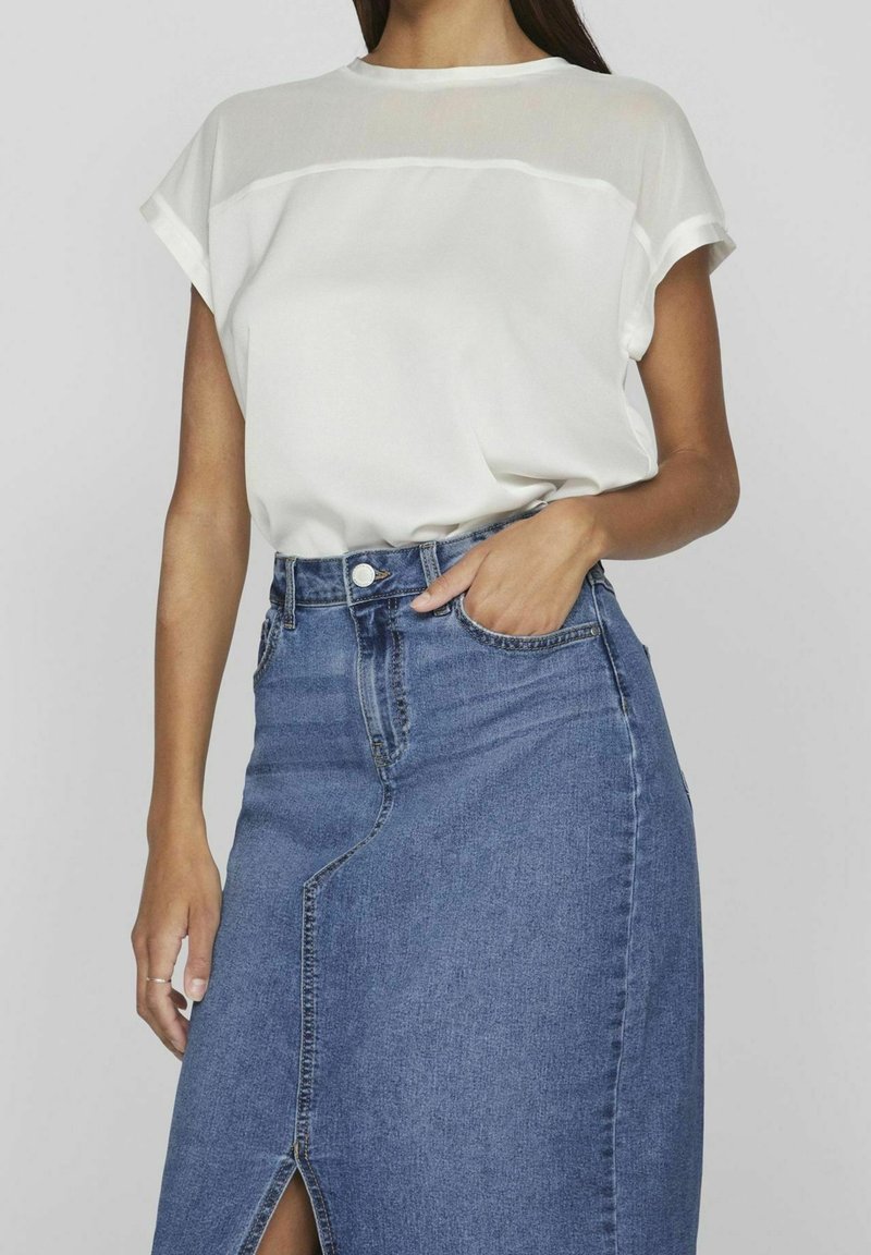 Femme portant un haut blanc à manches courtes rentré dans une jupe en denim bleue taille haute avec une fente devant et des poches.