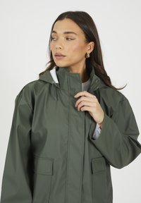 Veste imperméable vert olive avec un col montant, une capuche ajustable et deux poches frontales. Présente une texture lisse et une doublure contrastante.