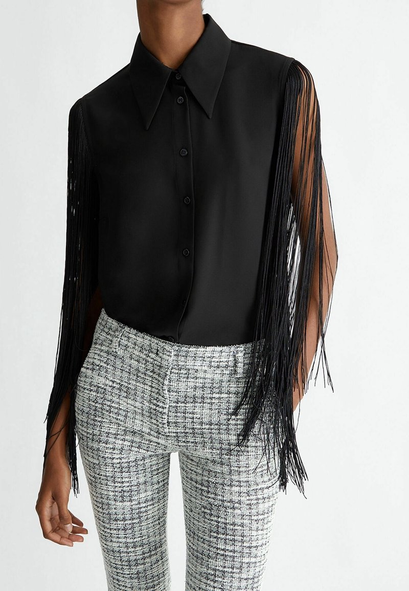 Blouse noire sans manches boutonnée avec de longues manches à franges, associée à un pantalon à carreaux noir et blanc taille haute.