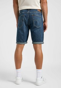 Shorts de denim en lavado oscuro con dobladillos enrollados, que presentan una etiqueta de cuero en la parte trasera, dos bolsillos en la parte posterior y detalles de costura amarilla.