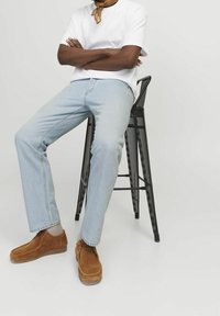 Zapatos de ante marrón con un detalle de costura, jeans de mezclilla azul claro y una camiseta blanca. Sentado en un taburete de metal negro perforado.