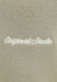 Svetlo béžová pletená látka s bielym vyšívaným textom „Originals Studio“ v kurzíve.