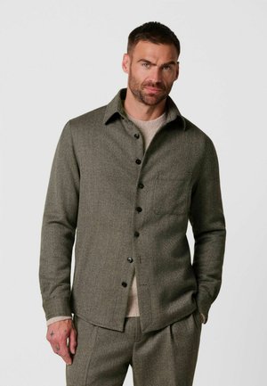 Uomo con capelli corti e barba che indossa una camicia grigia abbottonata sopra un maglione beige e pantaloni grigi coordinati, in piedi con una mano in tasca.