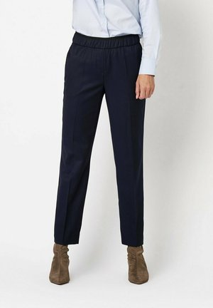 Dunkelblaue, gerade geschnittene Hose mit elastischem Bund, getragen zu einem hellblauen Hemd und braunen Wildleder-Stiefeletten vor weißem Hintergrund.