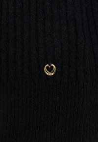 Svart ribbat stickat tyg har ett litet guldhjärtformat emblem. Texturen verkar mjuk och bekväm, vilket förbättrar dess visuella tilltalande.