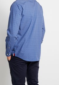 Blauwe gebloemde shirt met een knoopkraag, lange mouwen en een gebogen zoom. Bevat kleine witte geometrische prints en een contrasterend zijlabel.