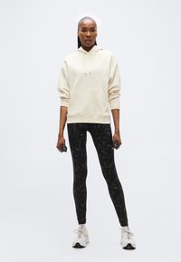 Γυναίκα που φοράει κρεμ φούτερ με κουκούλα και μαύρο κολάν με σχέδια, κρατώντας βαράκια 1 κιλού, στέκεται μπροστά σε λευκό φόντο.