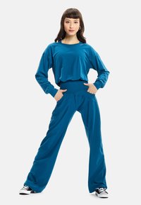 Set di abbigliamento attivo color teal composto da una maglietta lunga e ampia a maniche lunghe e pantaloni a gamba larga, realizzati in un tessuto morbido e liscio con un sottile dettaglio del logo.